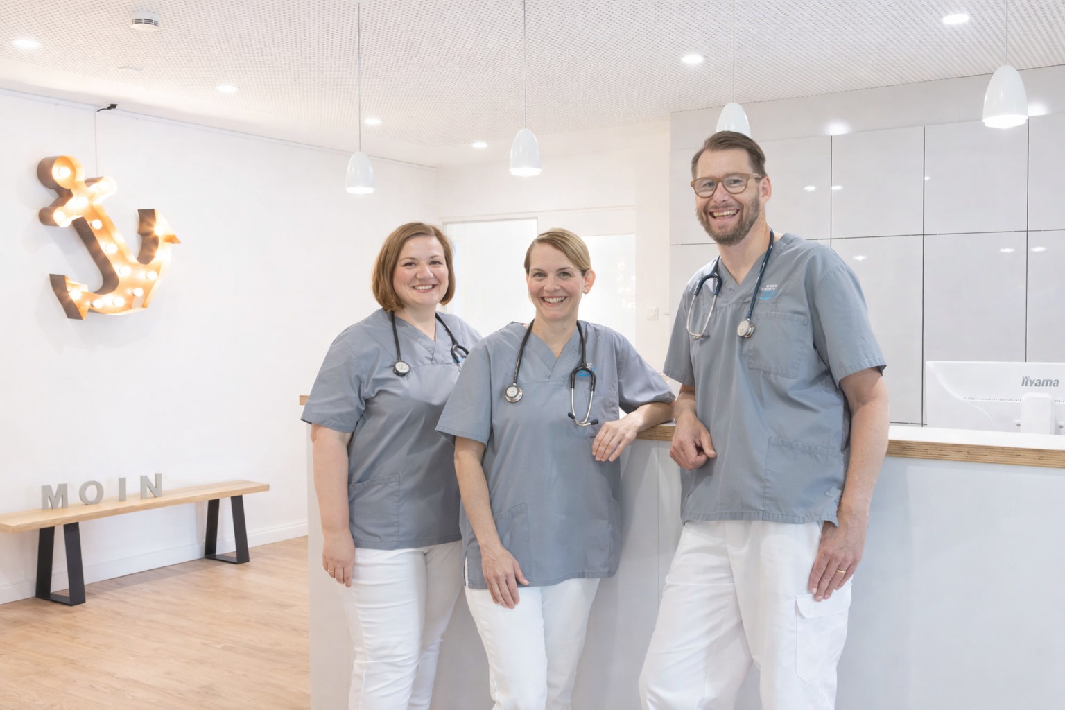 Vier lächelnde Ärztinnen und Ärzte in grauen Kasacks und weißen Hosen mit Stethoskopen um den Hals stehen gemeinsam am Empfangstresen der Gemeinschaftspraxis-Husum