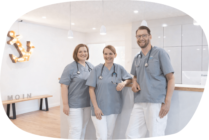 Vier lächelnde Ärztinnen und Ärzte in grauen Kasacks und weißen Hosen mit Stethoskopen um den Hals stehen gemeinsam am Empfangstresen der Gemeinschaftspraxis-Husum